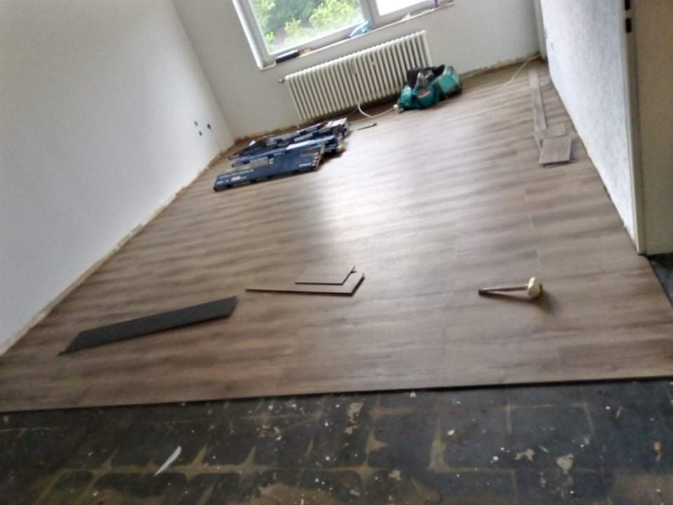 Fragment pokoju z częściowo ułożoną podłogą z paneli w odcieniach brązu, widoczne narzędzia i paczki paneli, stare podłoże w dolnej części kadru.