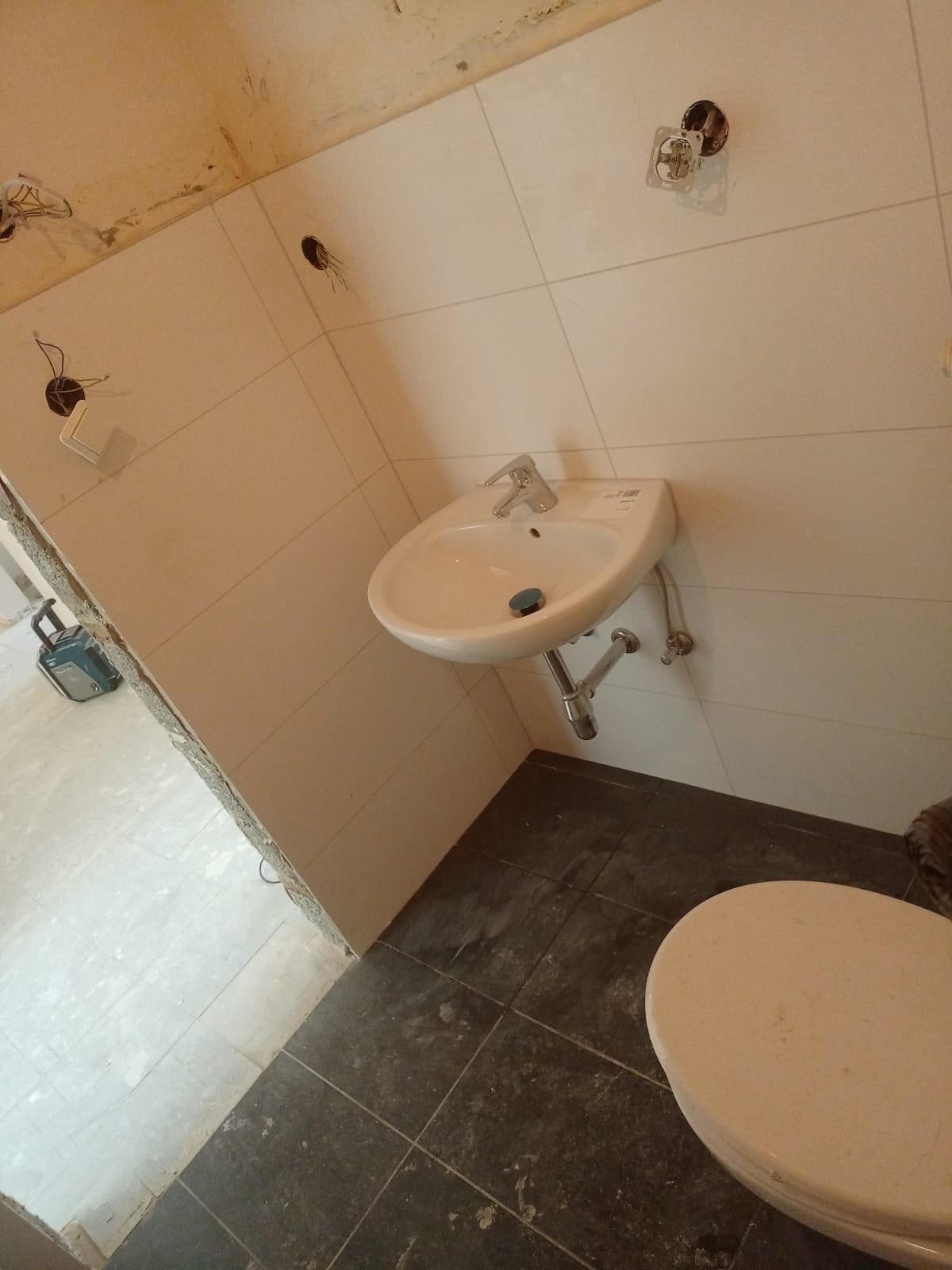 Łazienka w trakcie remontu: białe płytki ścienne, umywalka z baterią, częściowo ułożone ciemne płytki podłogowe, widoczne kable i puszki elektryczne bez osprzętu, narzędzie w tle