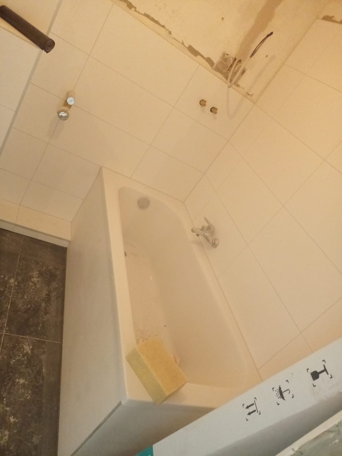 Niedokończona łazienka z nową wanną, białe płytki na ścianach, instalacja wodna widoczna na suficie, gąbka w wannie, widoczne przygotowania do dalszych prac wykończeniowych.