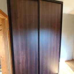 Meble na wymiar - Paweł Piórkowski - Dwudrzwiowa szafa przesuwna z ciemnego drewna z czarną ramą, widoczna na tle jasnych ścian i podłogi, z fragmentem ościeżnicy drzwi po lewej stronie.