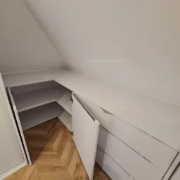 Biała szafa wnękowa na poddaszu ze skośnym sufitem i otwartymi drzwiami, ukazująca półki. Parkiet w jodełkę na podłodze. Minimalistyczny design.