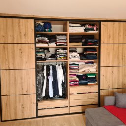 Meble na wymiar - Paweł Piórkowski - Jasna szafa przesuwna z drewnianymi frontami i czarnymi ramami, wypełniona ubraniami, widoczna z boku szara sofa z czerwoną narzutą. Realizacja.