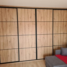 Meble na wymiar - Paweł Piórkowski - Duża, drewniana szafa przesuwna z czarnymi ramami, podzielona na 12 paneli. Obok szara sofa z czerwoną narzutą i poduszką. Jasne panele podłogowe.