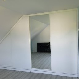 Meble na wymiar - Paweł Piórkowski - Biała szafa wnękowa z lustrzanymi drzwiami przesuwnymi, dopasowana do skosu poddasza w Tuchowie. Widoczna podłoga i fragment ściany w kolorze niebieskim.