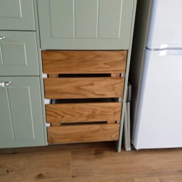 Meble na wymiar - Paweł Piórkowski - Fragment zabudowy kuchennej: pistacjowe szafki z drewnianymi szufladami w stylu rustykalnym, obok biała lodówka. Podłoga z paneli imitujących drewno.