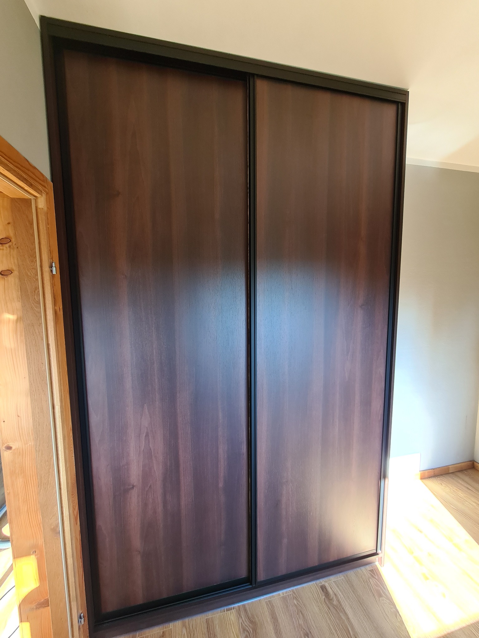 Dwudrzwiowa szafa przesuwna z ciemnego drewna z czarną ramą, widoczna na tle jasnych ścian i podłogi, z fragmentem ościeżnicy drzwi po lewej stronie.