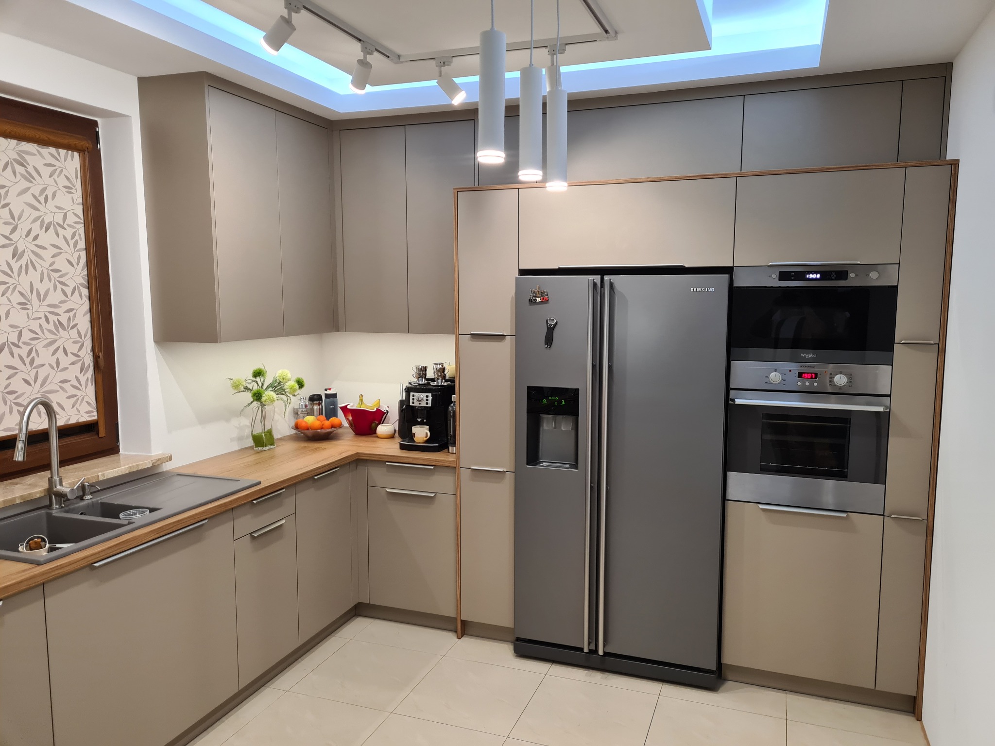 Nowoczesna kuchnia na wymiar w odcieniach beżu, z drewnianym blatem i sprzętem AGD w zabudowie. Widoczne oświetlenie punktowe i dekoracje na blacie.