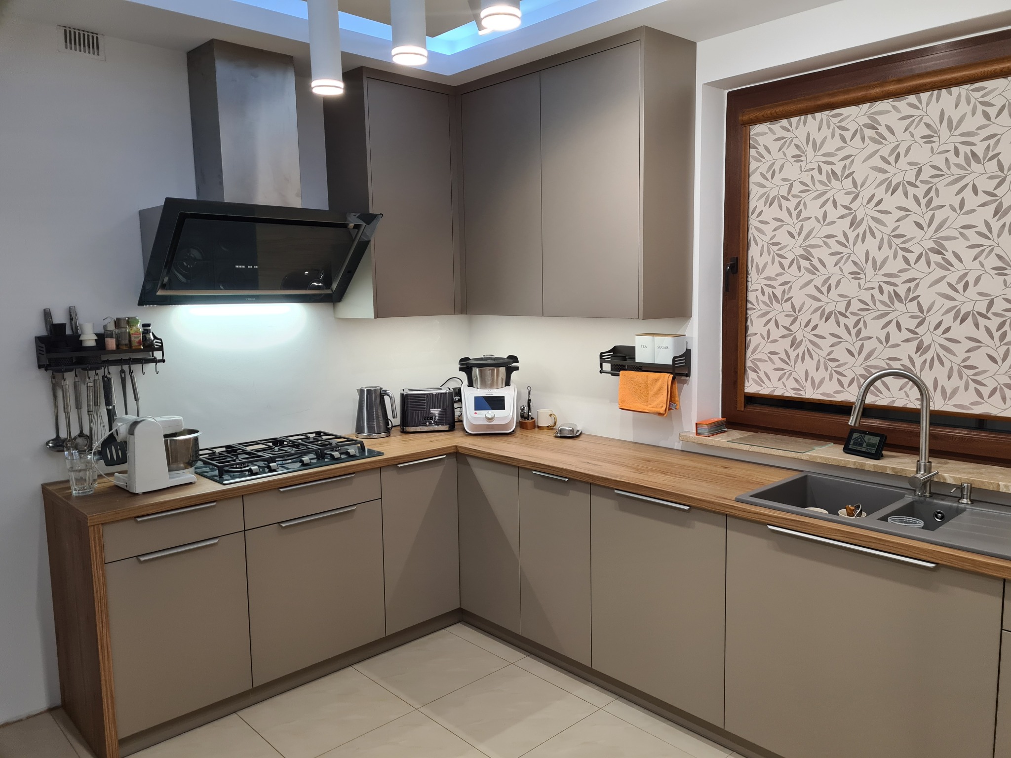 Jasna kuchnia z szarymi frontami, drewnianym blatem i oknem z roletą w roślinny wzór. Na blacie kuchenka gazowa, zlew i drobne sprzęty AGD.
