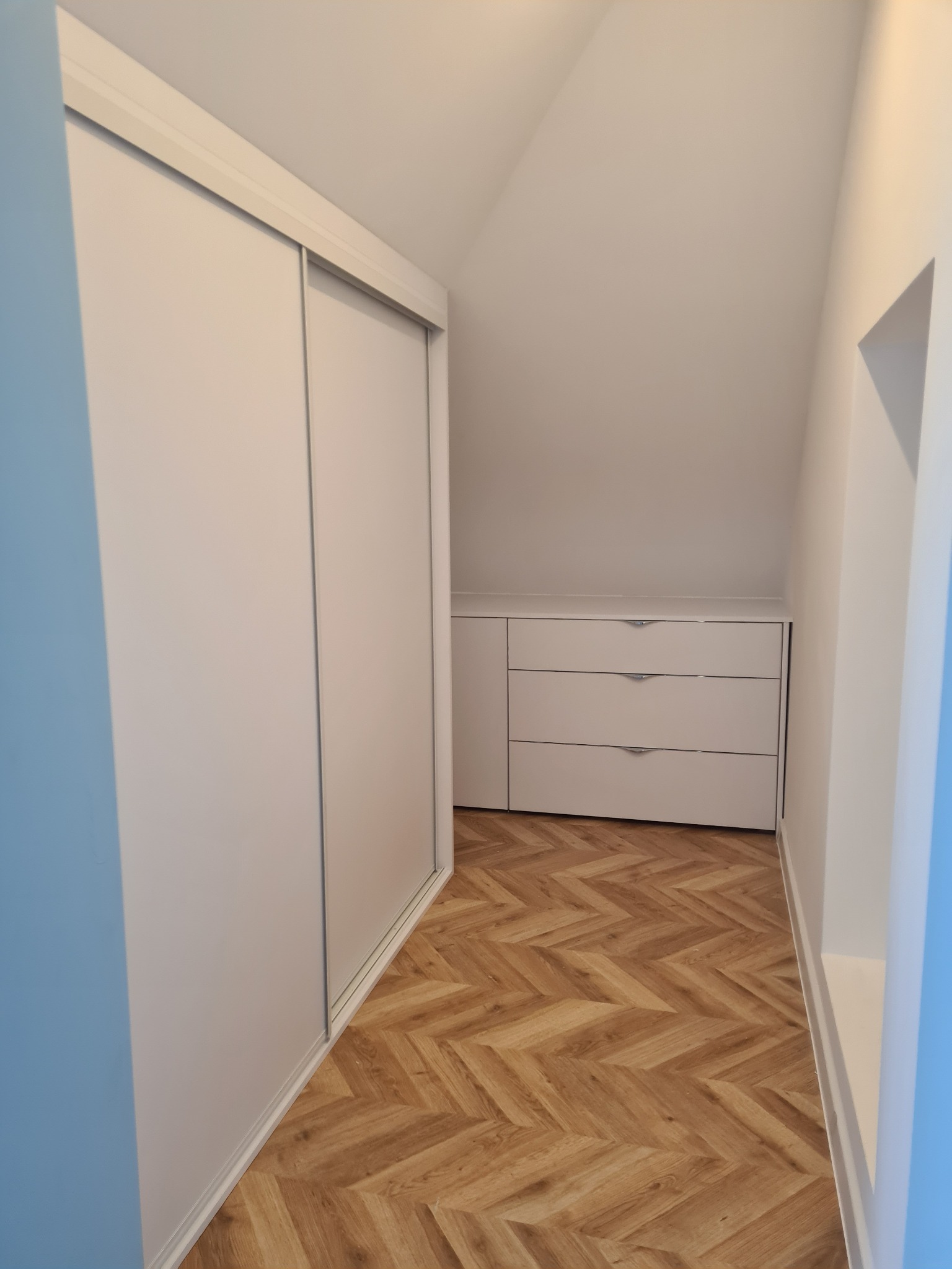 Biała szafa przesuwna i komoda z trzema szufladami wnękowe na poddaszu, parkiet w jodełkę. Funkcjonalne zagospodarowanie przestrzeni.