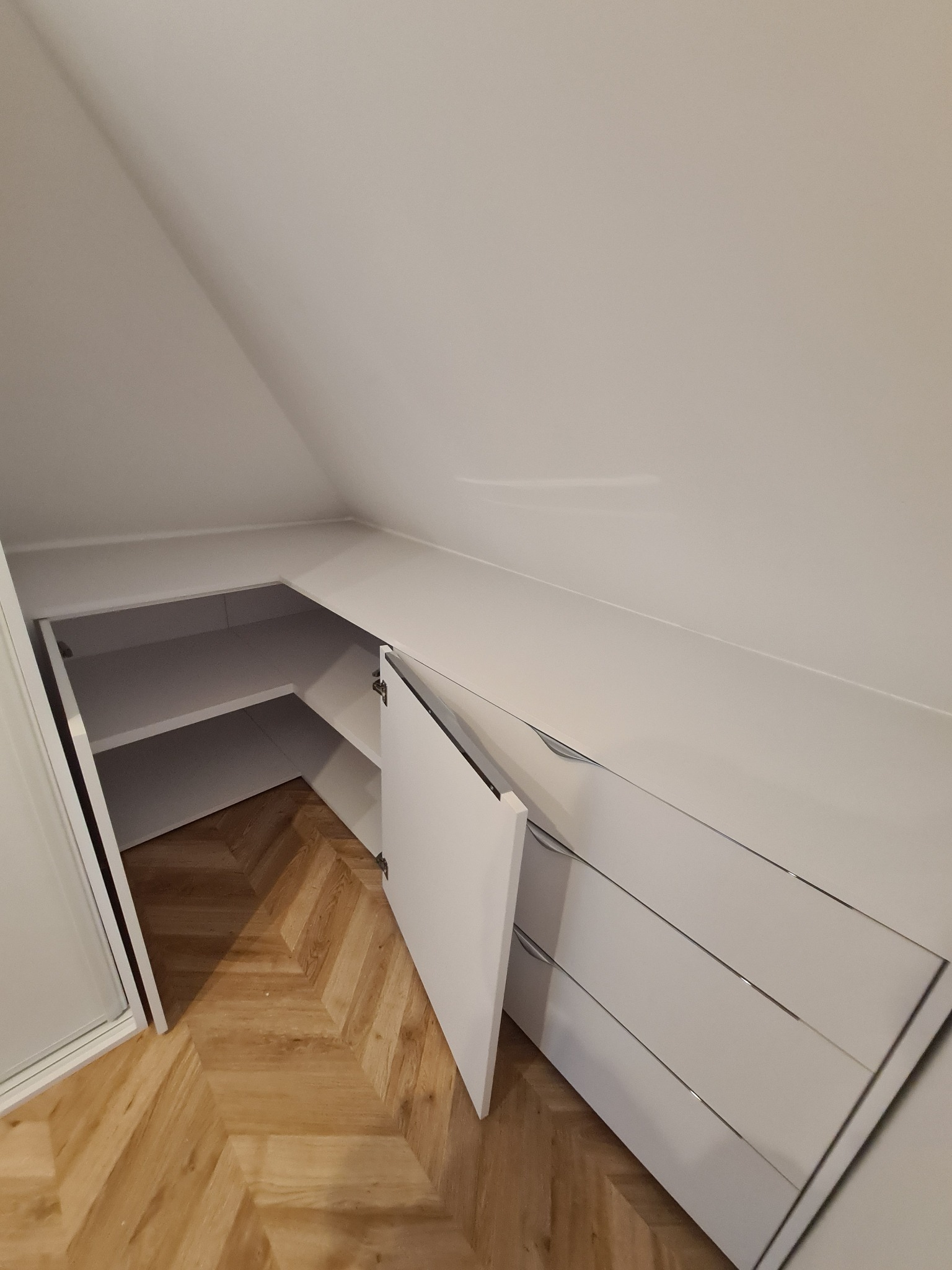 Biała szafa wnękowa na poddaszu ze skośnym sufitem i otwartymi drzwiami, ukazująca półki. Parkiet w jodełkę na podłodze. Minimalistyczny design.