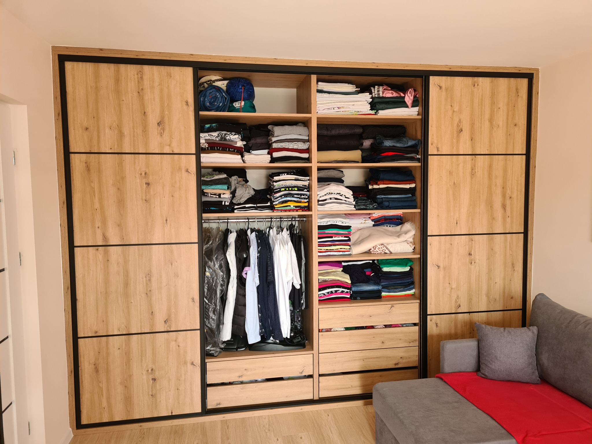 Jasna szafa przesuwna z drewnianymi frontami i czarnymi ramami, wypełniona ubraniami, widoczna z boku szara sofa z czerwoną narzutą. Realizacja.