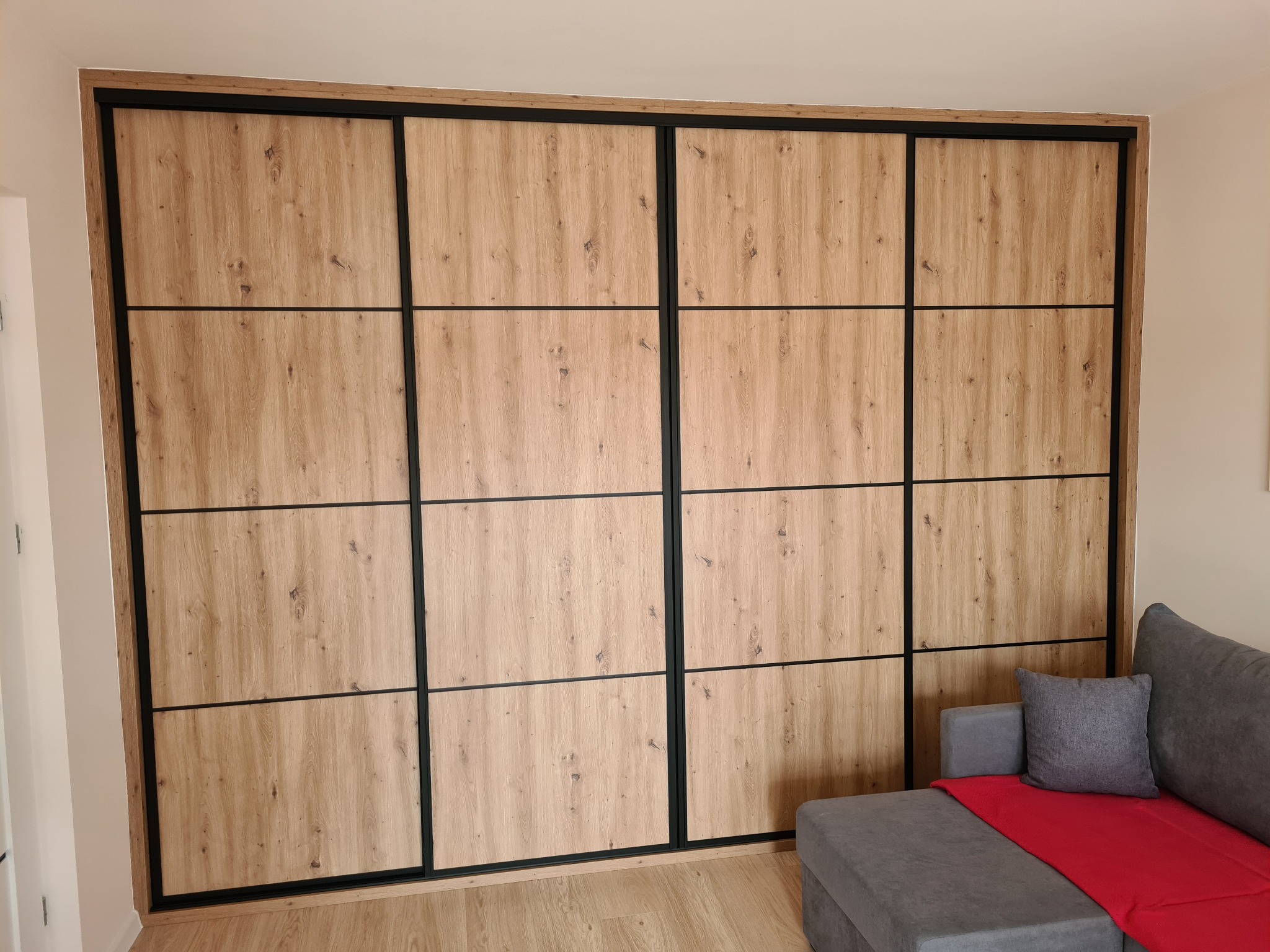Duża, drewniana szafa przesuwna z czarnymi ramami, podzielona na 12 paneli. Obok szara sofa z czerwoną narzutą i poduszką. Jasne panele podłogowe.