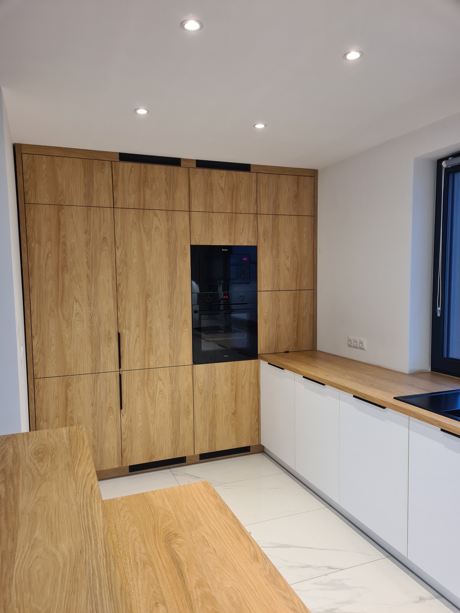 Nowoczesna kuchnia: drewniane szafki z czarnymi akcentami, białe dolne szafki i marmurowe płytki podłogowe. Uwagę przyciąga wbudowany piekarnik.