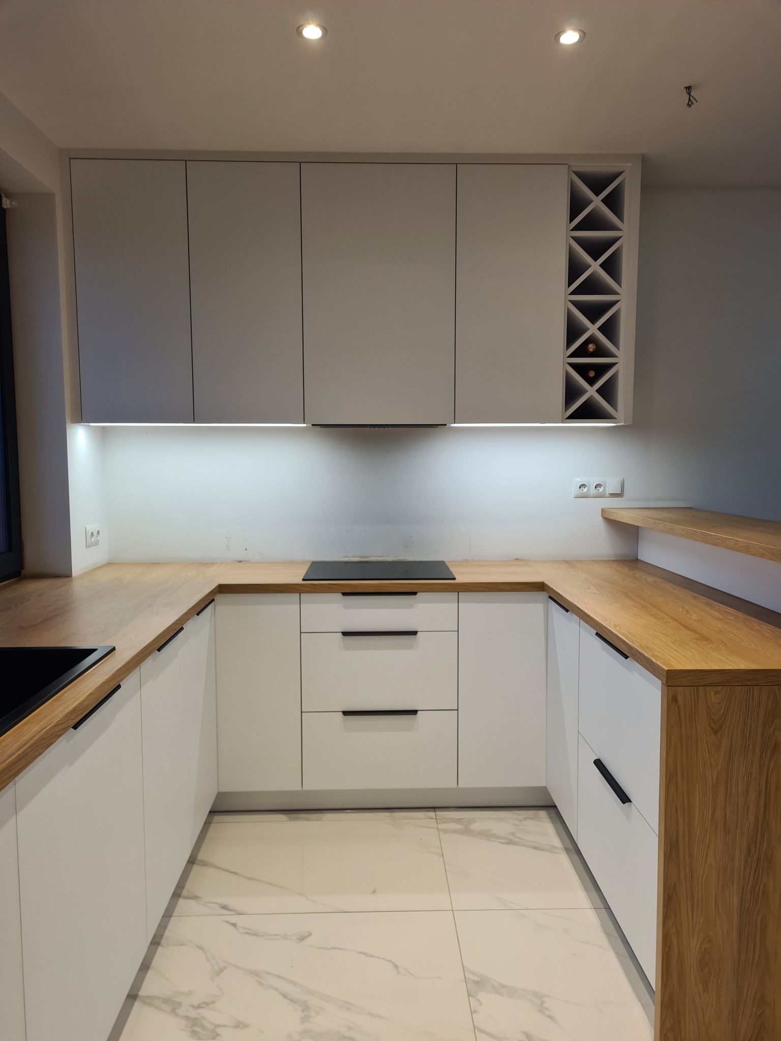 Nowoczesna, jasna kuchnia na wymiar z drewnianym blatem i wbudowaną półką na wino. Minimalistyczne uchwyty, białe fronty i marmurowe płytki podłogowe.