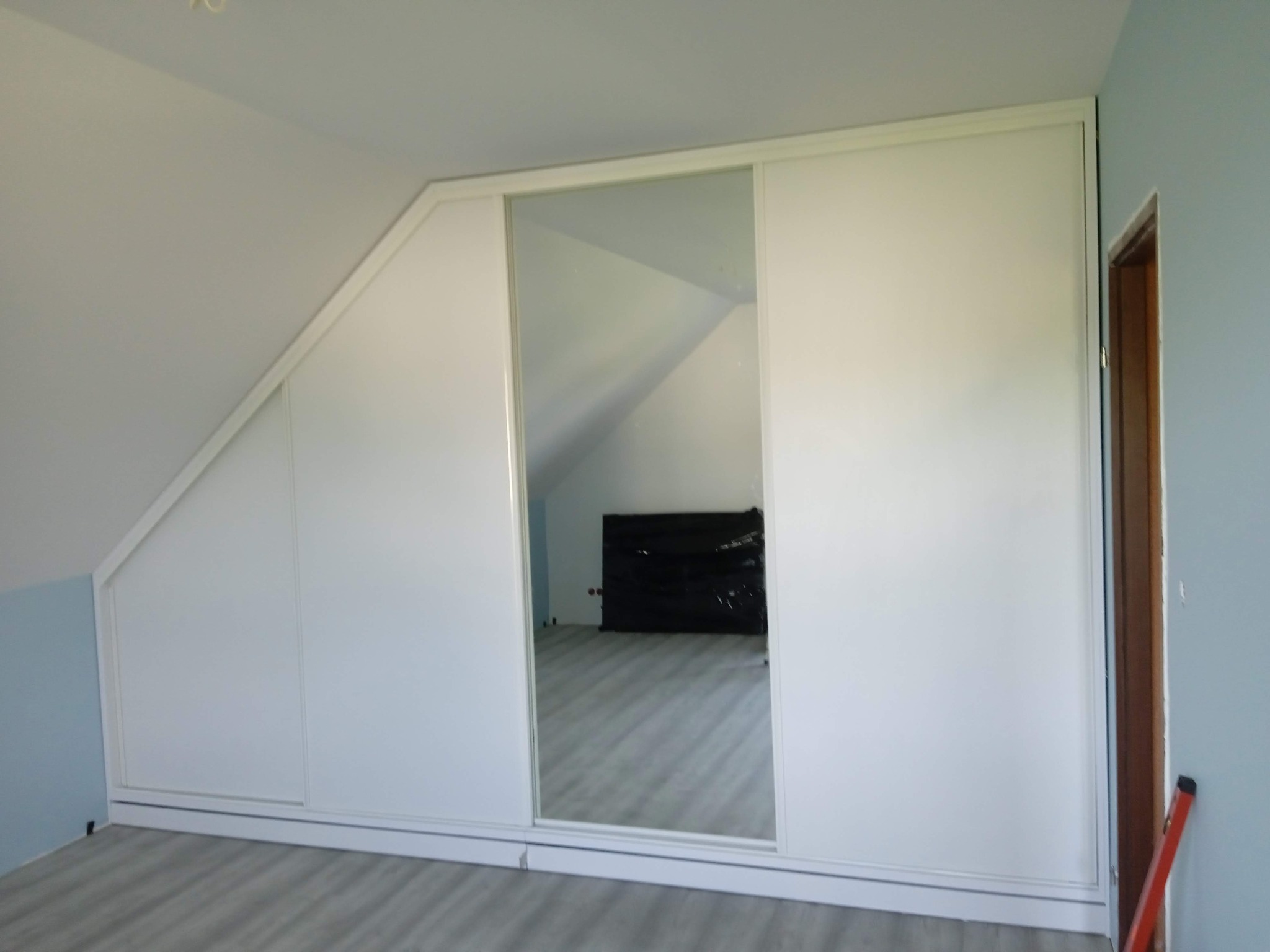 Biała szafa wnękowa z lustrzanymi drzwiami przesuwnymi, dopasowana do skosu poddasza w Tuchowie. Widoczna podłoga i fragment ściany w kolorze niebieskim.