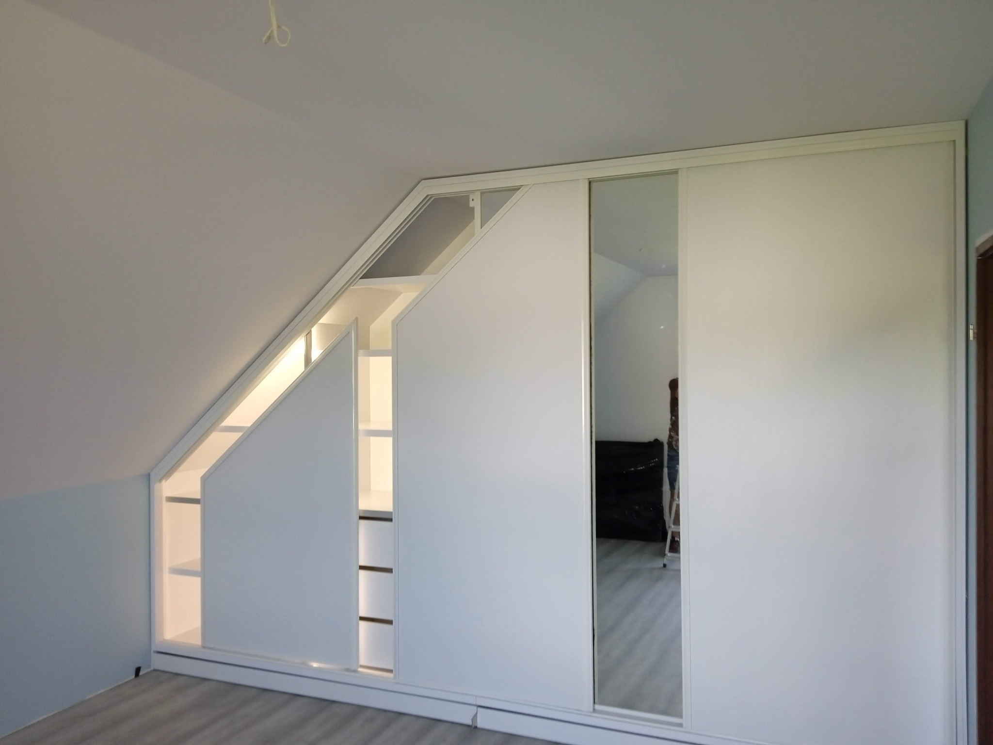 Biała szafa wnękowa na poddaszu ze skośnym frontem i przesuwnymi drzwiami, widoczne półki i oświetlenie. W lustrze odbija się drabina.