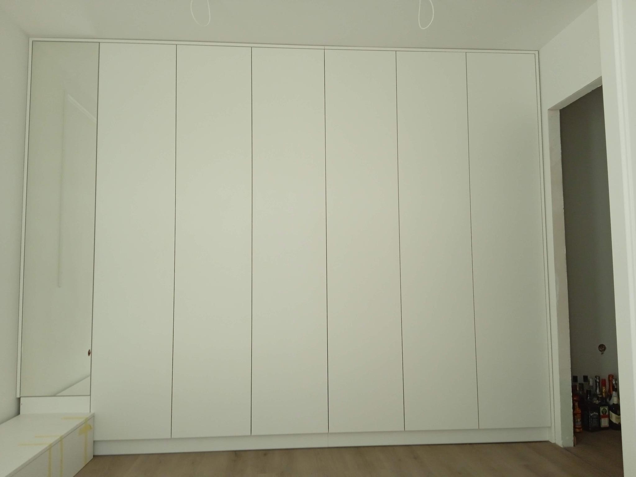 Biała szafa wnękowa z lustrem i szafką, minimalistyczny design, jasna podłoga. Uwagę przykuwają butelki widoczne w przejściu obok szafy.