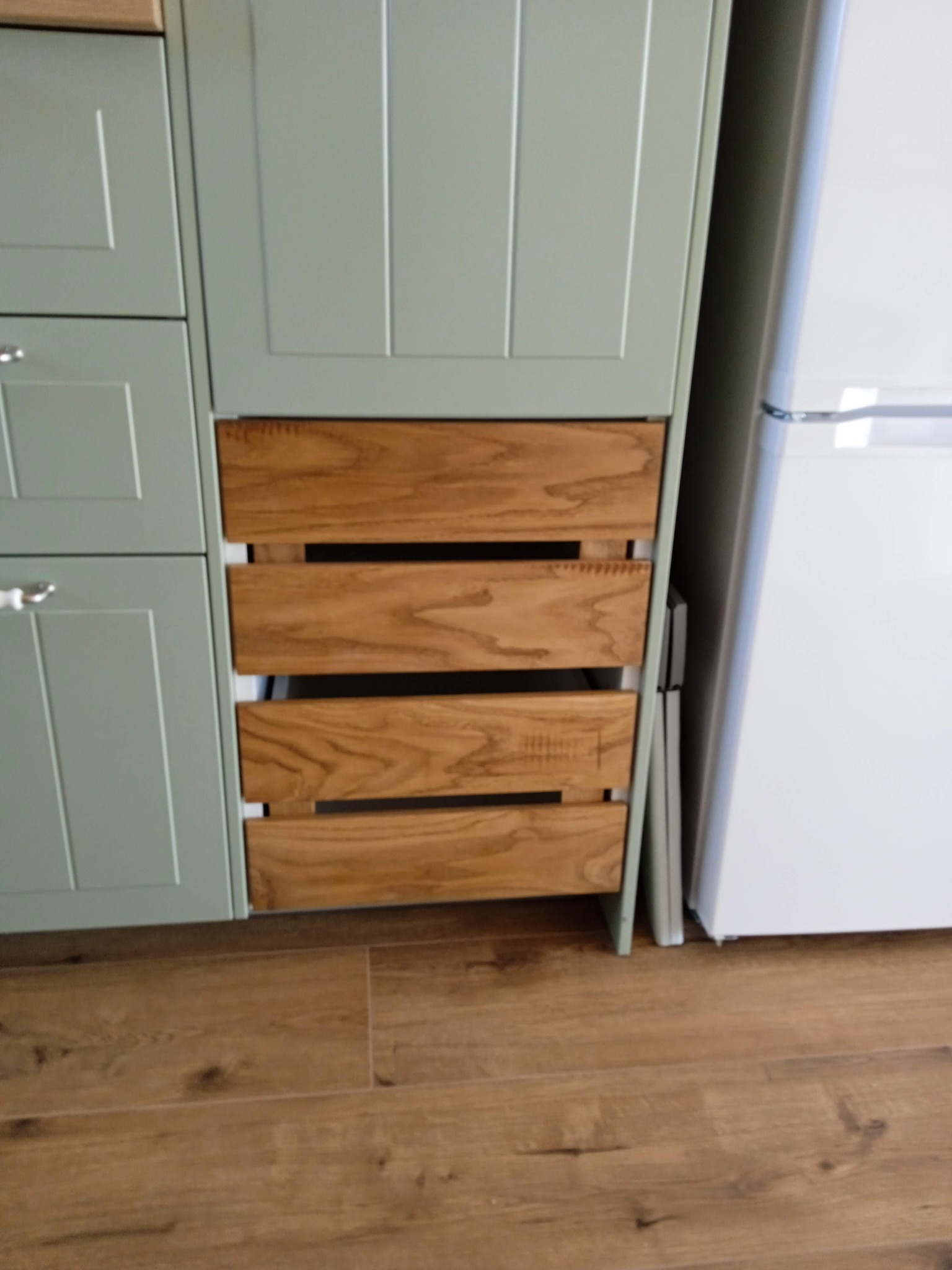 Fragment zabudowy kuchennej: pistacjowe szafki z drewnianymi szufladami w stylu rustykalnym, obok biała lodówka. Podłoga z paneli imitujących drewno.