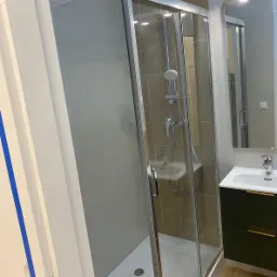 Nowoczesna kabina prysznicowa z przesuwnymi drzwiami, obok umywalka z szafką w odcieniach zieleni i złotymi uchwytami, wykończenie łazienki w szaro-beżowych płytkach.