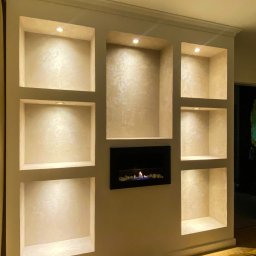 ToMaz-Bud - Ściana z wnękami oświetlonymi punktowo, wykończona dekoracyjnym tynkiem. W jednej z wnęk biokominek. Widoczny fragment łóżka z żółtą narzutą.