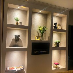 ToMaz-Bud - Ściana z wnękami oświetlonymi punktowo, dekorowanymi różnymi przedmiotami, w tym książką, kwiatami, rzeźbami i kominkiem elektrycznym. Dekoracyjne wykończenie ścian.