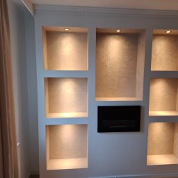 ToMaz-Bud - Ściana z wnękami oświetlonymi punktowo, wykończonymi w beżowym odcieniu, obok zasłony i czarny element dekoracyjny. Widoczne narzędzia malarskie.