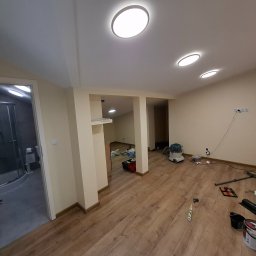 ToMaz-Bud - Przestronne wnętrze po remoncie z drewnianą podłogą, widok na łazienkę z kabiną prysznicową. Na podłodze widoczne narzędzia i materiały malarskie.