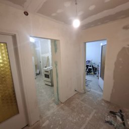 ToMaz-Bud - Wnętrze w trakcie remontu: widoczne ślady szpachlowania na ścianach i suficie, przygotowanie do malowania. W tle widoczny sprzęt i narzędzia remontowe.