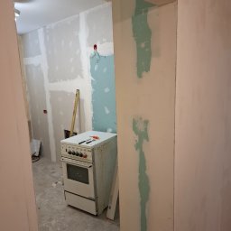ToMaz-Bud - Remont kuchni w trakcie: ściany z płyt karton-gips, częściowo zagruntowane, stara kuchenka i poziomica w tle. Widok z sąsiedniego pomieszczenia.