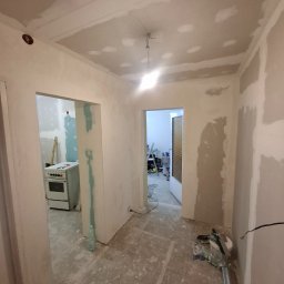 ToMaz-Bud - Remont mieszkania w trakcie: ściany i sufit po szpachlowaniu, widoczne narzędzia i przygotowania do dalszych prac wykończeniowych. W tle kuchnia z kuchenką.