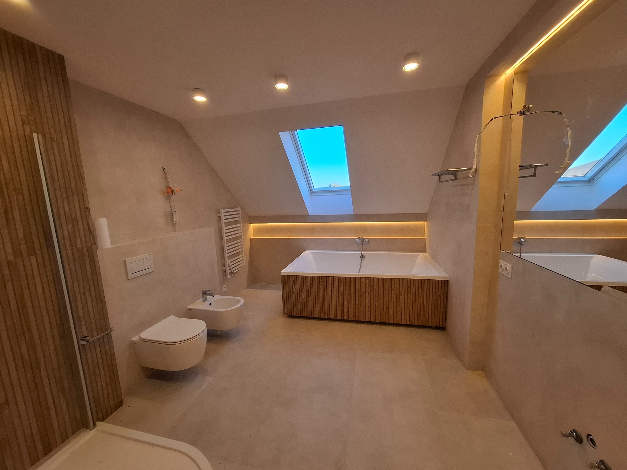 Jasna łazienka na poddaszu z oknem dachowym, drewnianą obudową wanny i beżowymi płytkami. Umywalka, toaleta i grzejnik drabinkowy dopełniają wystrój.