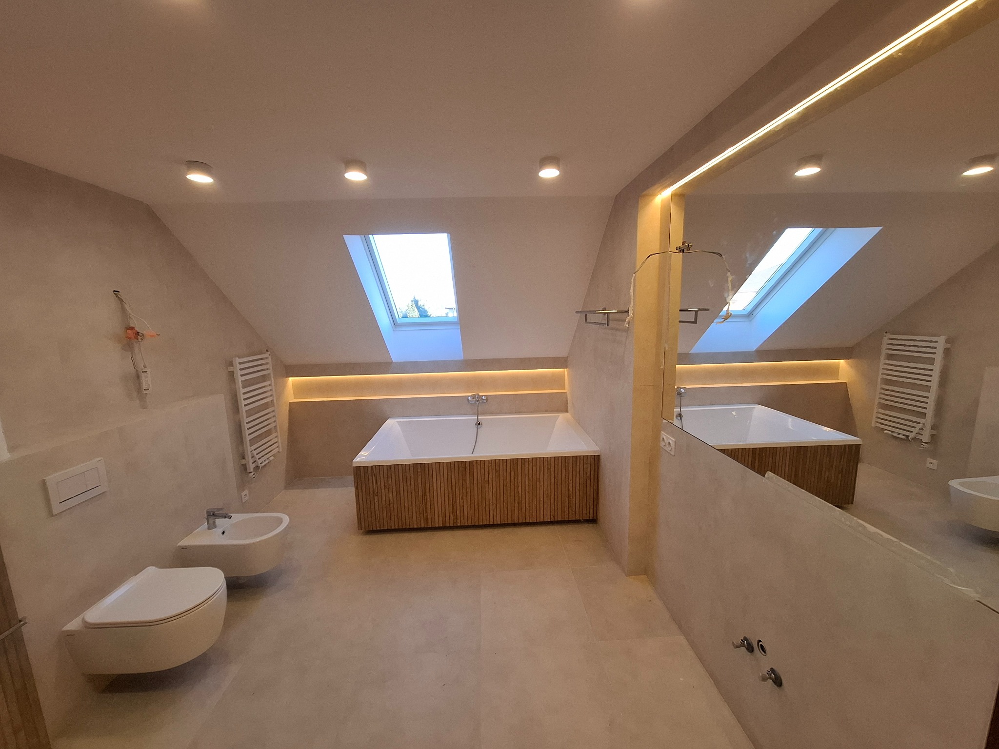 Jasna łazienka na poddaszu z wanną obudowaną drewnem, bidetem i dużym lustrem z oświetleniem LED. Widoczne okno dachowe i grzejnik drabinkowy. Całość utrzymana w beżowej tonacji.