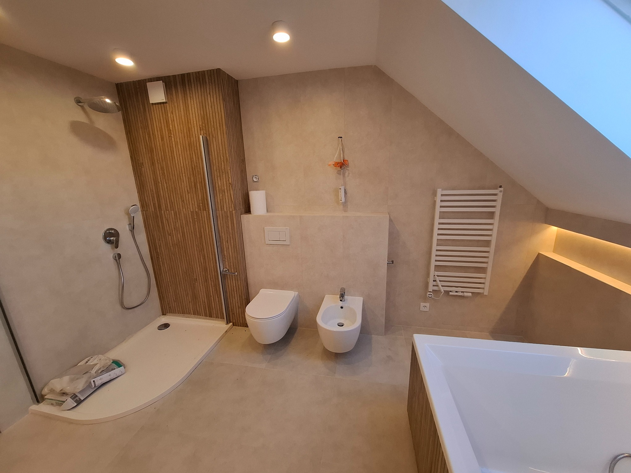 Wykończona łazienka na poddaszu z kabiną prysznicową, toaletą, bidetem i grzejnikiem drabinkowym. Użyto beżowych płytek i drewnopodobnych paneli. Widoczne skosy.
