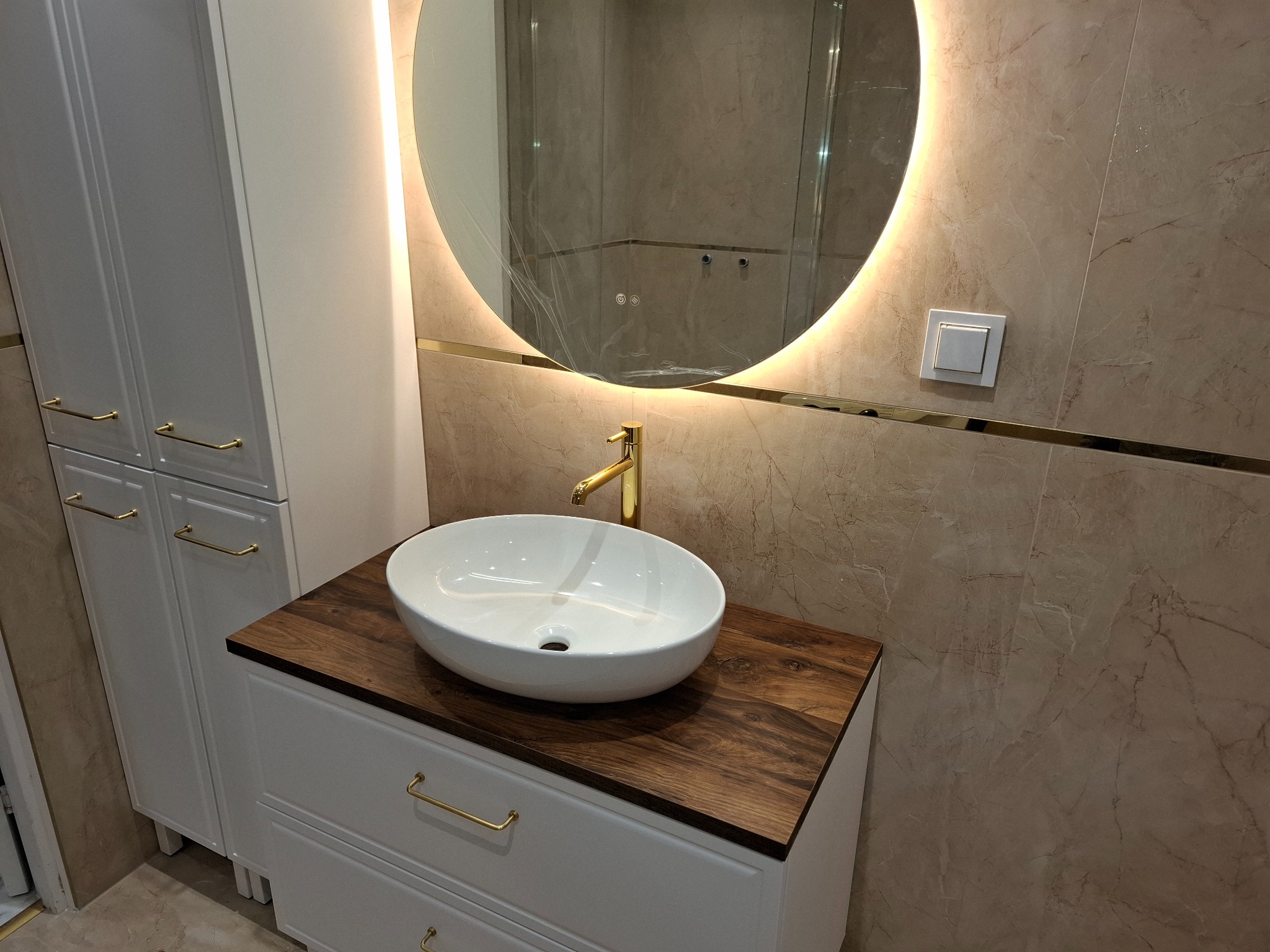 Nowoczesna łazienka: biała szafka z drewnianym blatem, okrągłe lustro z podświetleniem LED i złota armatura na tle marmurowych płytek.