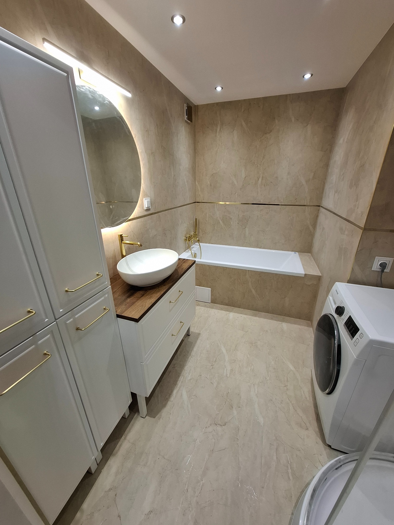Wykończona łazienka w beżach z białą armaturą: umywalka na drewnianym blacie, szafki z złotymi uchwytami, wanna i pralka. Całość oświetlona punktowo i listwą LED.