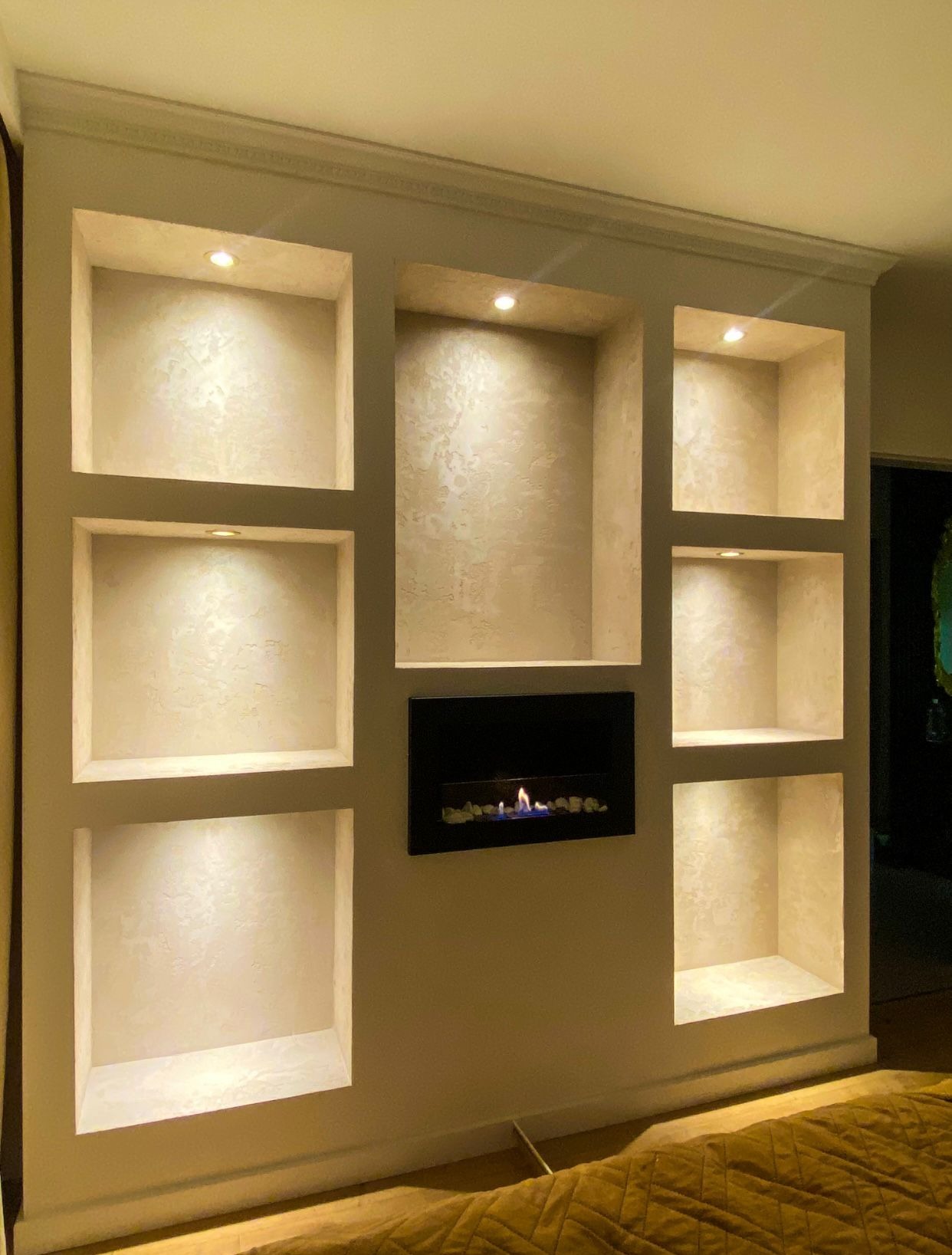 Ściana z wnękami oświetlonymi punktowo, wykończona dekoracyjnym tynkiem. W jednej z wnęk biokominek. Widoczny fragment łóżka z żółtą narzutą.