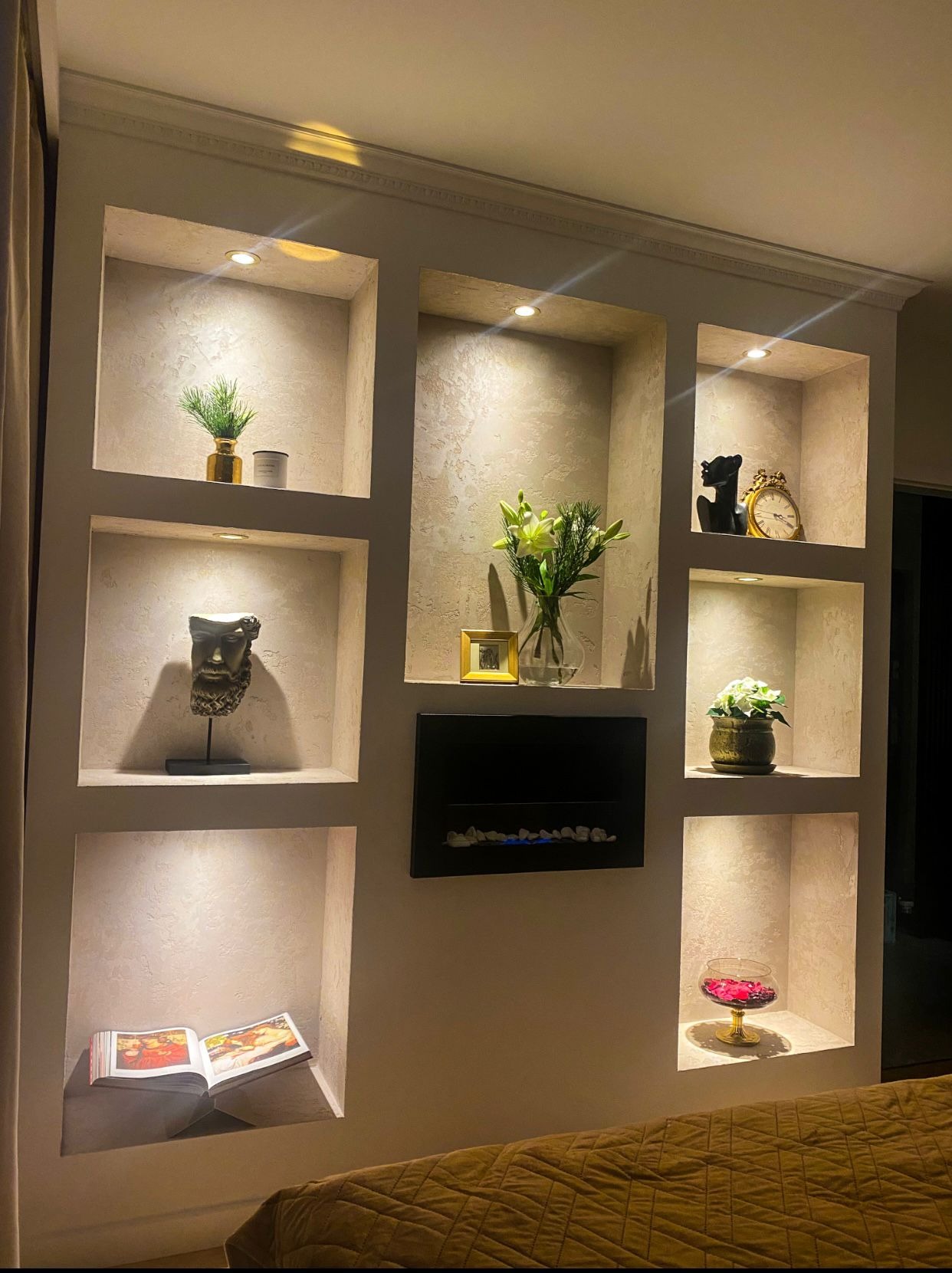 Ściana z wnękami oświetlonymi punktowo, dekorowanymi różnymi przedmiotami, w tym książką, kwiatami, rzeźbami i kominkiem elektrycznym. Dekoracyjne wykończenie ścian.