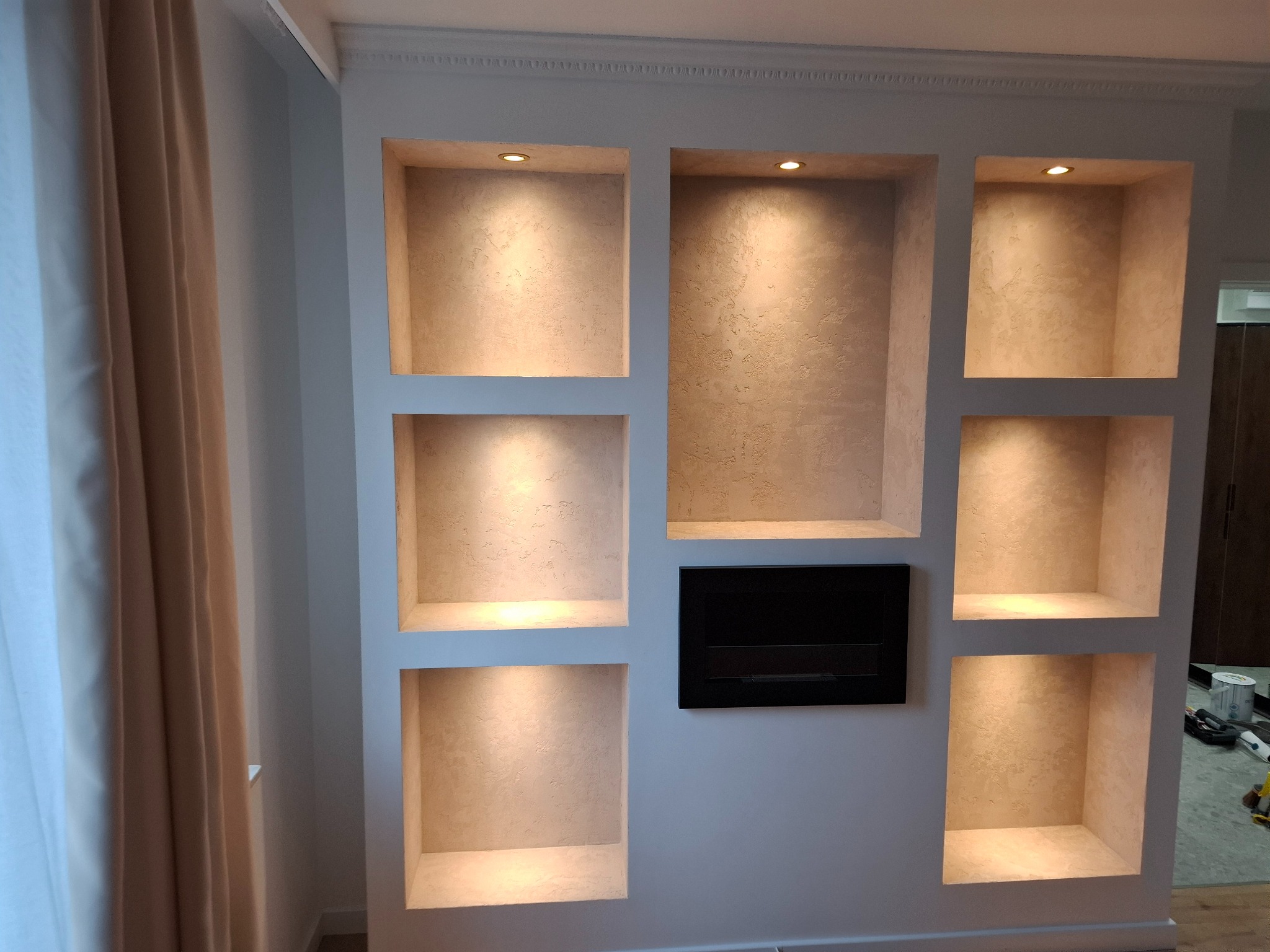 Ściana z wnękami oświetlonymi punktowo, wykończonymi w beżowym odcieniu, obok zasłony i czarny element dekoracyjny. Widoczne narzędzia malarskie.