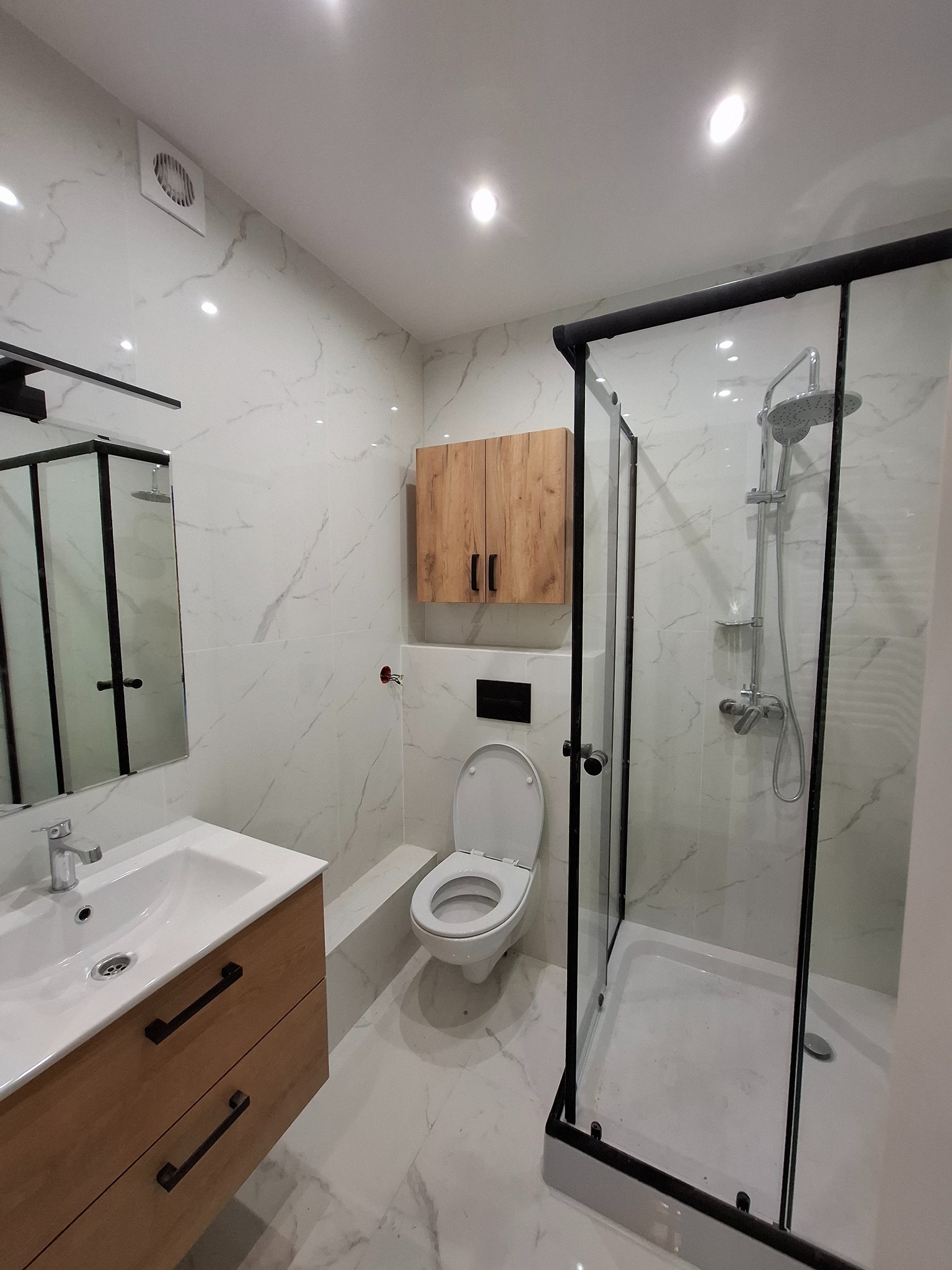 Nowoczesna łazienka z kabiną prysznicową typu walk-in, umywalką z szafką z drewnianym frontem i wiszącą szafką nad toaletą. Białe płytki imitujące marmur.