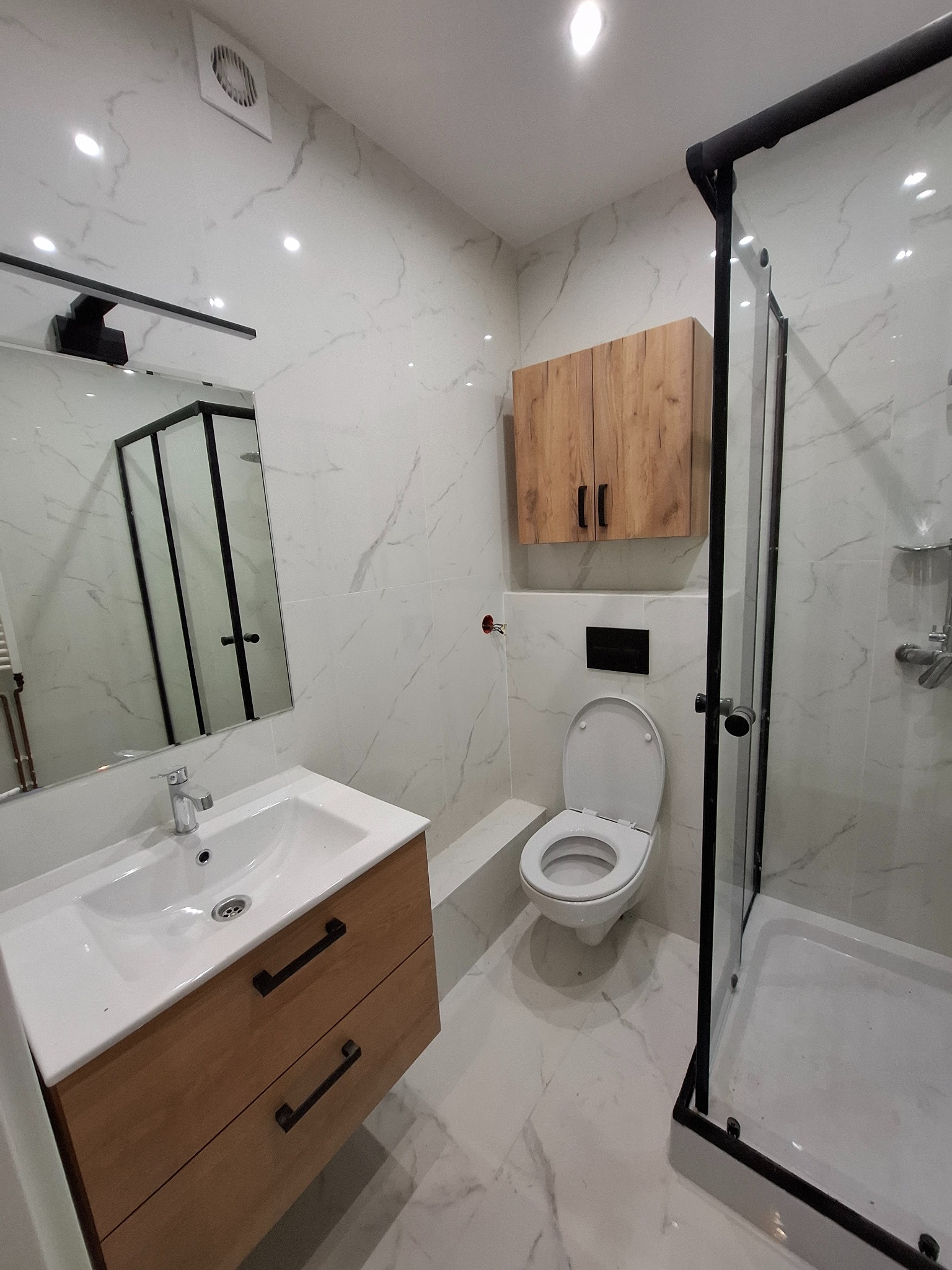 Nowoczesna łazienka z marmurowymi płytkami, drewnianą szafką pod umywalką i kabiną prysznicową z czarnymi profilami. Uwagę zwraca minimalistyczna zabudowa toalety.