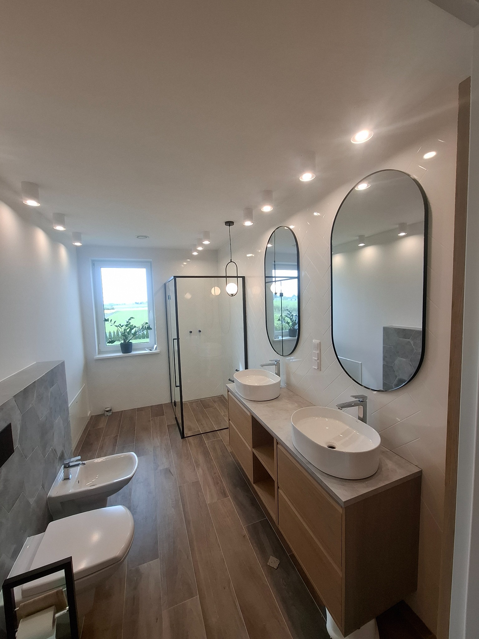 Nowoczesna łazienka z drewnianą podłogą, owalnymi lustrami i kabiną prysznicową w czarnej ramie. Umywalki nablatowe i biała armatura.