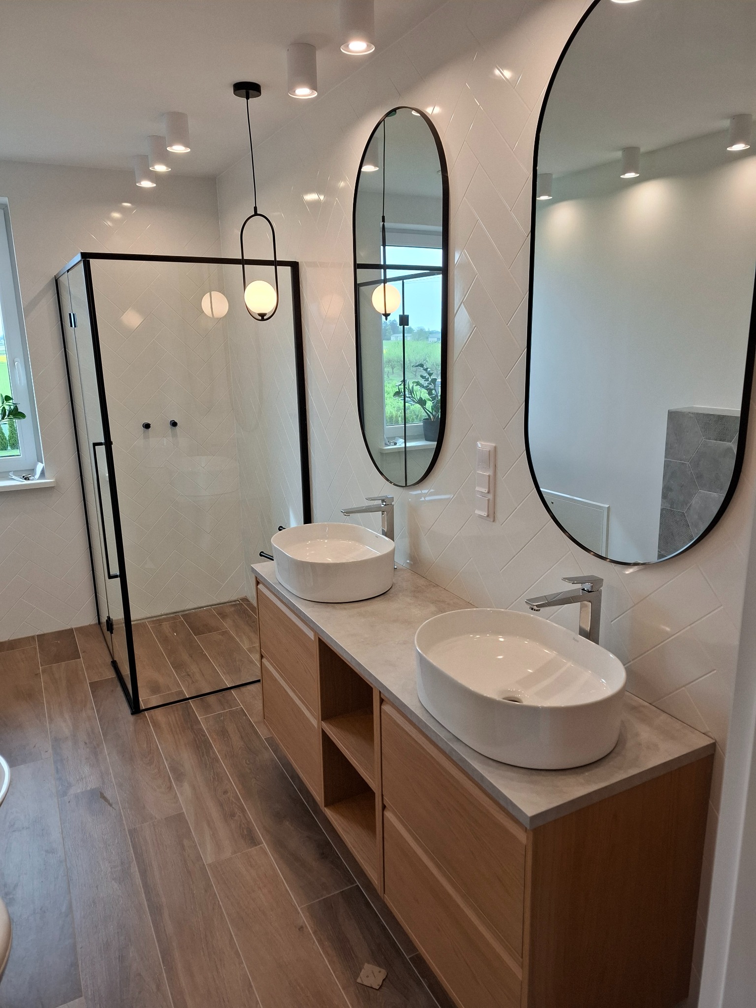 Nowoczesna łazienka z kabiną prysznicową typu walk-in, podwójną umywalką nablatową i owalnymi lustrami. Białe płytki ułożone w jodełkę i drewnopodobna podłoga.
