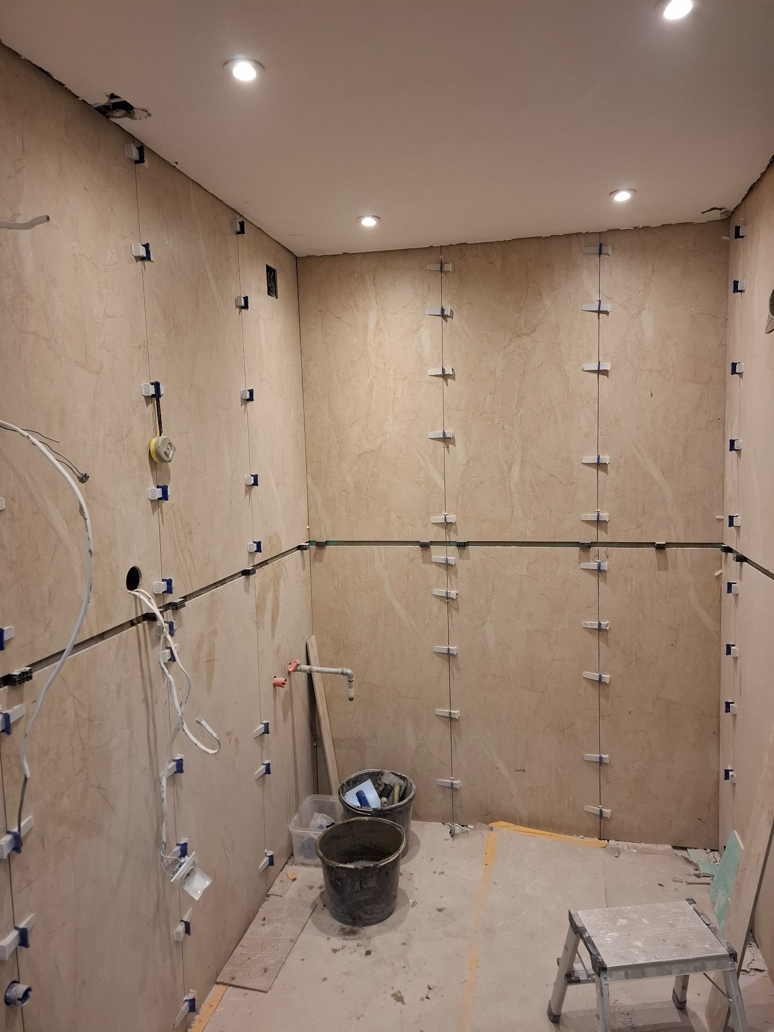 Wnętrze łazienki w trakcie remontu: beżowe płytki na ścianach z niebieskimi krzyżykami dystansowymi, widoczne kable i narzędzia, przygotowane do fugowania.