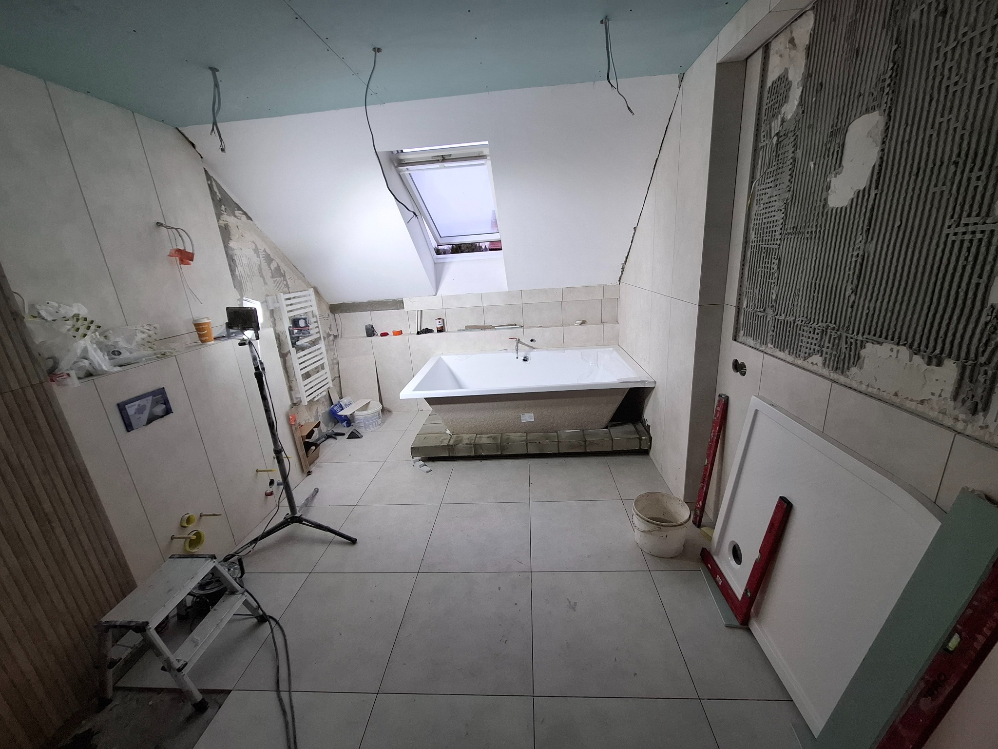 Remont łazienki na poddaszu: biała wanna pod oknem dachowym, częściowo położone płytki ścienne, narzędzia i materiały budowlane w trakcie prac wykończeniowych.
