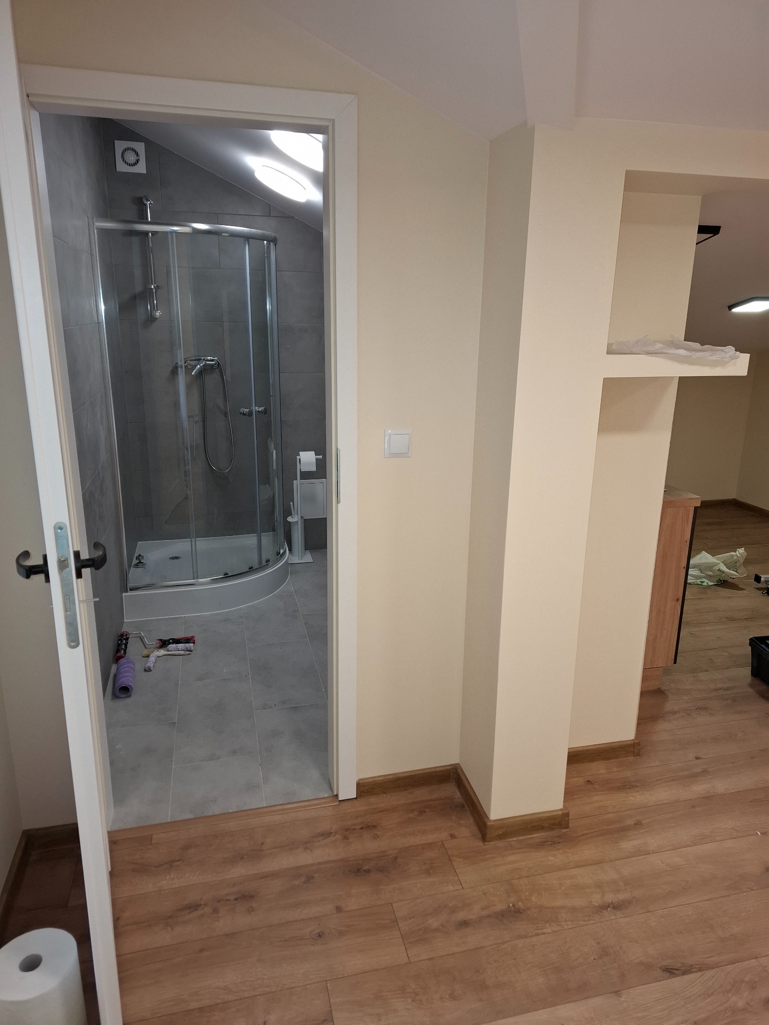 Wyremontowana łazienka z kabiną prysznicową i szarymi płytkami, widoczna z korytarza z drewnianą podłogą. Na podłodze w łazience leżą wałki do malowania.