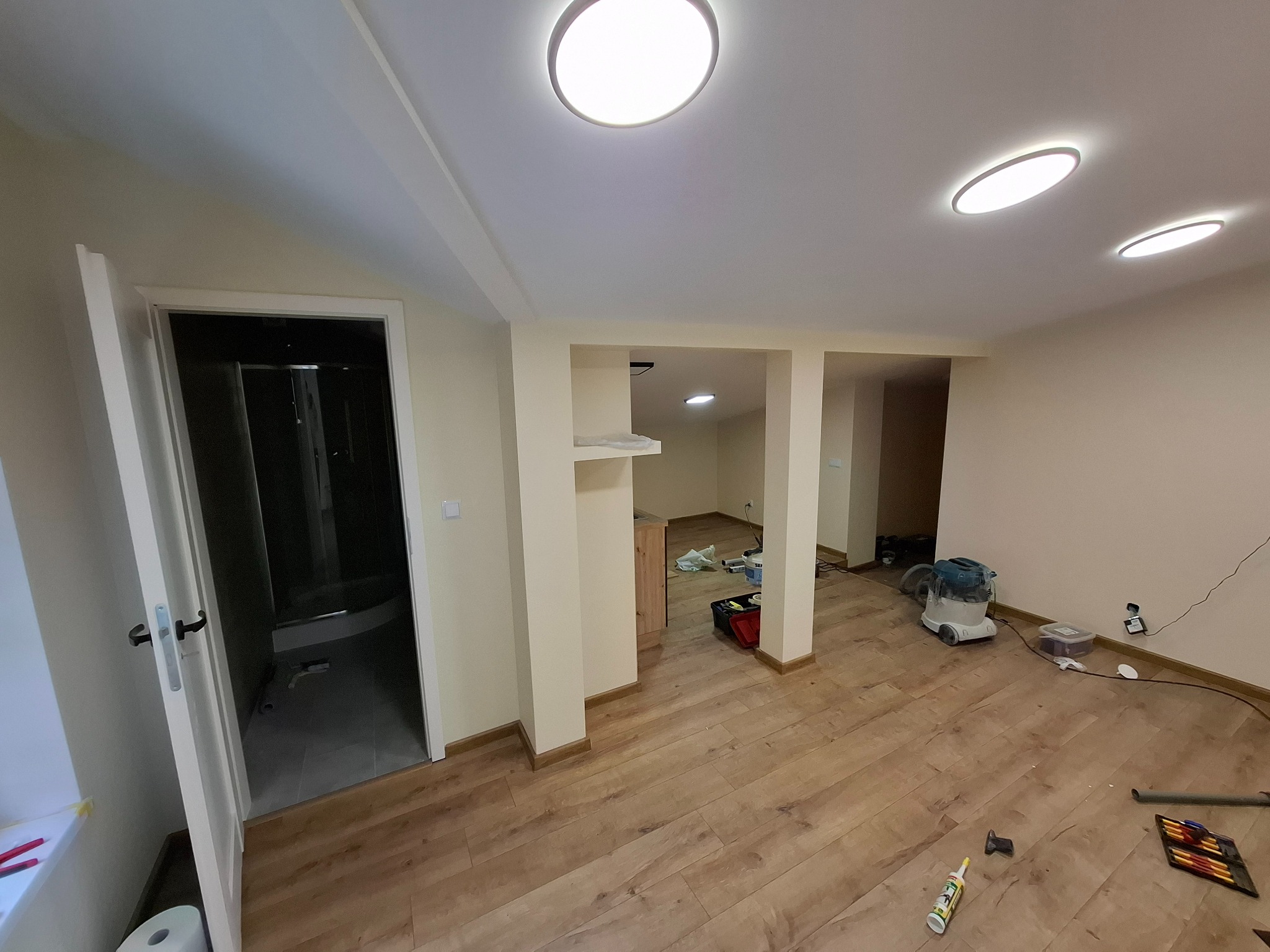 Jasne wnętrze po remoncie z drewnianą podłogą, otwartą łazienką z kabiną prysznicową i narzędziami na podłodze. Widoczne oświetlenie sufitowe i świeżo pomalowane ściany.