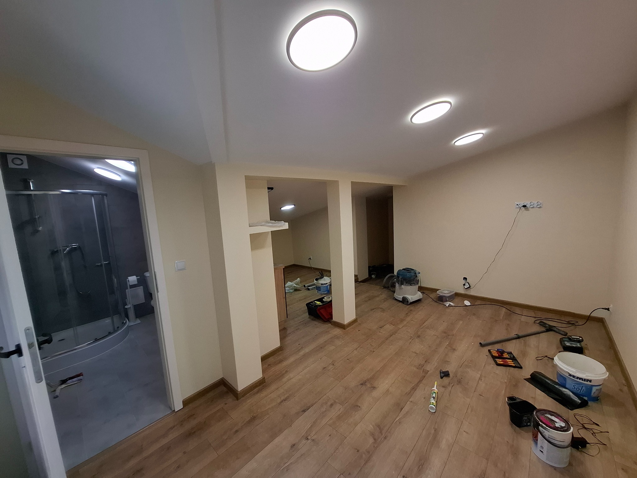 Przestronne wnętrze po remoncie z drewnianą podłogą, widok na łazienkę z kabiną prysznicową. Na podłodze widoczne narzędzia i materiały malarskie.