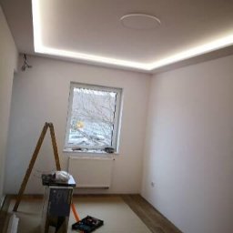 Tomasz Banaś - Wykończenia Wnętrz - Pokój z białymi ścianami i sufitem z oświetleniem LED, drabina, narzędzia i materiały na podłodze, okno z widokiem na drzewa.