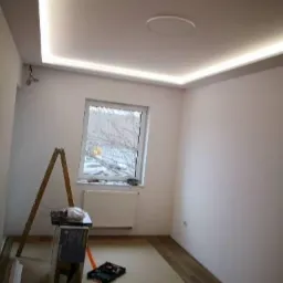 Pokój z białymi ścianami i sufitem z oświetleniem LED, drabina, narzędzia i materiały na podłodze, okno z widokiem na drzewa.