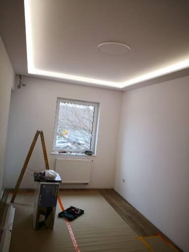 Pokój z białymi ścianami i sufitem z oświetleniem LED, drabina, narzędzia i materiały na podłodze, okno z widokiem na drzewa.